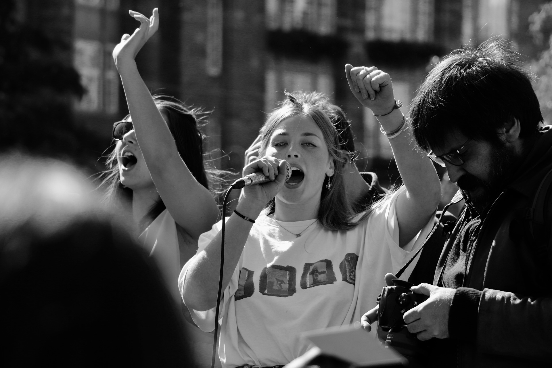 a young woman standing in a protesting crowd with a microphone held up to her mouth and fist in the air 
